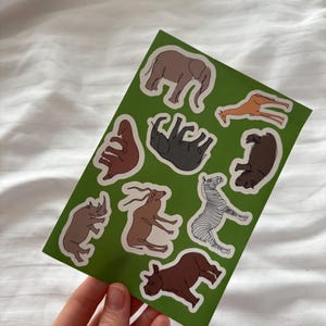 May include: A sheet of animal stickers featuring illustrations of an elephant, giraffe, zebra, rhino, and hippo. The stickers have a white border and are on a green background. The stickers are held by a hand.