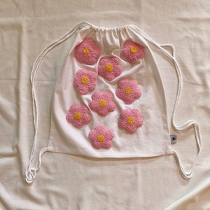Op de afbeelding: Een witte trekkoordtas met geborduurde roze en gele bloemen. De tas heeft een wit trekkoord en is gemaakt van een zachte, natuurlijke stof.