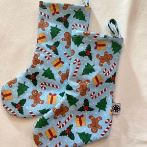 May include: Two Christmas stockings with a light blue background and a repeating pattern of gingerbread men, candy canes, wrapped gifts, and Christmas trees. Each stocking has a loop for hanging.