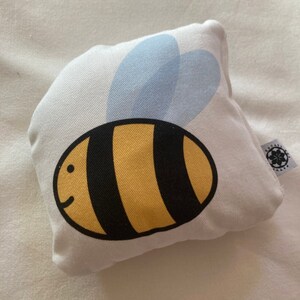 May include: A white pillow with a yellow and black bee design. The bee has a blue and white wing.