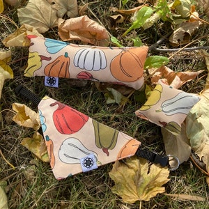 Puede incluir: Un pañuelo para mascotas, un juguete rectangular y un pequeño juguete cuadrado, todos de color beige y con un estampado de calabazas coloridas. El pañuelo tiene una correa negra ajustable. Los juguetes tienen una pequeña etiqueta blanca con un logotipo negro.