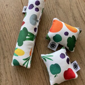Puede incluir: Tres almohadas de tela coloridas con fondo blanco y una variedad de diseños de frutas y verduras. Las almohadas son rectangulares y cuadradas. Las almohadas tienen un logotipo en blanco y negro que dice "Play for Fun" en ellas.