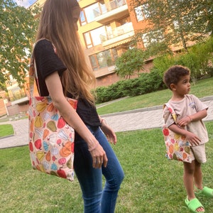 Può includere: Due borse tote con un motivo a zucche colorate. Una borsa è portata da una persona che indossa un top nero e jeans blu. L'altra borsa è portata da un bambino che indossa una maglietta e pantaloncini beige. Entrambi sono sull'erba verde.