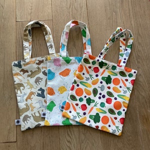 Könnte beinhalten: Drei kleine Tragetaschen mit verschiedenen Mustern. Eine Tasche hat einen beigen Hintergrund mit verschiedenen Tierillustrationen. Eine andere hat einen weißen Hintergrund mit bunten Zeichentricktieren. Die dritte Tasche zeigt einen weißen Hintergrund mit bunten Gemüseillustrationen.