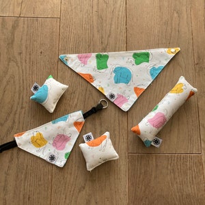 Puede incluir: Un conjunto de accesorios para mascotas con una tela blanca estampada con ilustraciones de elefantes de colores. El conjunto incluye dos bandanas triangulares, dos pequeñas almohadas cuadradas y un juguete cilíndrico largo.