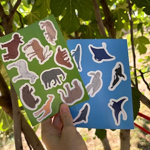 May include: Two sticker sheets featuring animal illustrations. One sheet is green and has land animals like a zebra, elephant, and giraffe. The other sheet is blue and has sea creatures like a manta ray and orca. The stickers have white outlines.