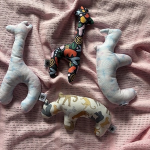 May include: Four plush stuffed animal toys on a pink knitted blanket. The toys are a giraffe with a black, white, orange, and red pattern, a giraffe with a blue and white pattern, a rabbit with a blue and white pattern, and a brown and white animal with a pattern of various animals.