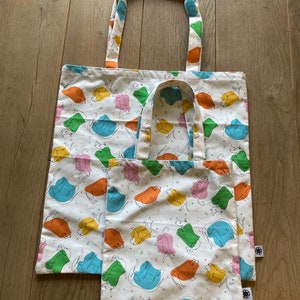 Könnte beinhalten: Drei weiße Tragetaschen mit einem bunten Katzenmuster. Die Katzen sind schwarz umrandet und mit Orange, Gelb, Grün, Blau und Rosa gefüllt. Die Taschen liegen auf einer Holzoberfläche.