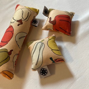 Puede incluir: Tres juguetes para gatos de varias formas. Los juguetes son de color beige con un patrón repetido de pimientos rojos, amarillos y verdes. Cada juguete tiene una pequeña etiqueta con un diseño floral y texto.