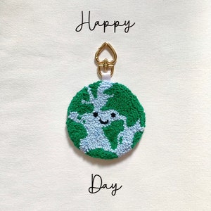 Pode incluir: Um chaveiro em forma de Terra verde e azul com um rosto sorridente. O chaveiro tem um fecho dourado em forma de coração e as palavras "Happy Day" estão escritas abaixo.