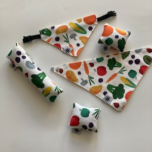 Puede incluir: Un conjunto de accesorios para mascotas, que incluye pañuelos y pequeñas almohadas, con un estampado de verduras colorido. La tela blanca está adornada con ilustraciones de zanahorias, brócoli, tomates y otras verduras. El pañuelo tiene un collar negro ajustable.