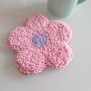 May include: A pink and purple crocheted flower coaster. The coaster is shaped like a flower with five petals and a center circle.