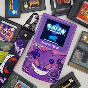 Puede incluir: Una consola de videojuegos portátil Game Boy Color de color morado con un cartucho de juego Pokemon Edición Cristal insertado. La consola tiene un diseño morado y rosa con un personaje Pokemon con una gran sonrisa. La consola está rodeada de otros cartuchos de juego.