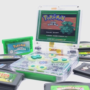 Può includere: Una console Game Boy Advance SP trasparente con dettagli verdi, che mostra "Pokémon Edición Verde Hoja" sullo schermo. Sono visibili diverse cartucce di gioco, tra cui "Pokémon Esmeralda". I pulsanti e il D-pad della console sono verdi.