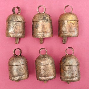 May include: Six antique metal bells with a rustic, aged appearance. Each bell has a loop at the top and a wooden clapper. The bells are arranged on a pink background, showcasing their unique textures and tones.