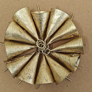 May include: A golden metal decorative piece featuring a circular arrangement of cone-shaped bells. Each bell has a textured surface and is attached to a central spiral. The bells are arranged around a central point with small metal rods extending outwards.