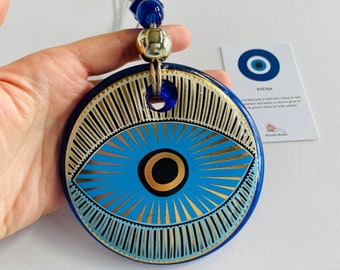 Op de afbeelding: Een blauwe en gouden wanddecoratie in de vorm van een boze oog met een blauwe en gouden kralenketting. Het boze oog is een symbool van bescherming tegen pech en jaloezie. De tekst op de kaart luidt "Evil Eye" en "Het wordt algemeen aangenomen dat het ons beschermt tegen ongelukken en schade, daarom geven we het aan degenen die we echt bewonderen."