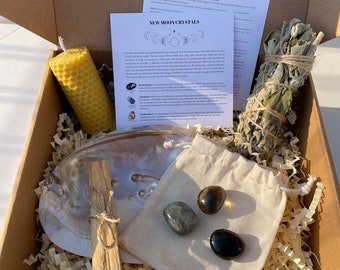 May include: A New Moon Crystals kit in a cardboard box. The kit includes a yellow honeycomb candle, a white shell, a bundle of sage, a palo santo stick, a small bag with three crystals, and informational cards.