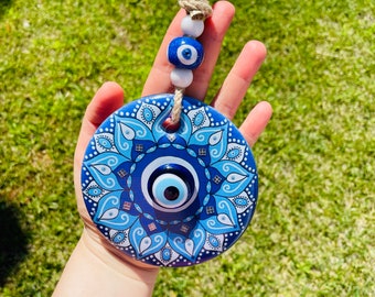 Op de afbeelding: Een blauwe en witte keramische wanddecoratie met een boze oog en een mandala-ontwerp. Het boze oog is omgeven door een blauw en wit bloemenpatroon.
