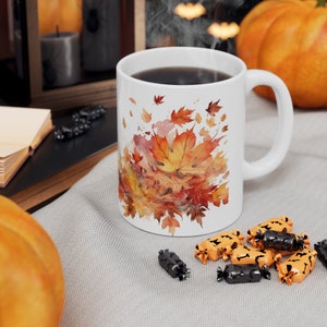 Pode incluir: Caneca de cerâmica branca com um desenho de aquarela de folhas de outono em tons de laranja, amarelo e vermelho. A caneca está cheia de café e vapor está subindo do topo.