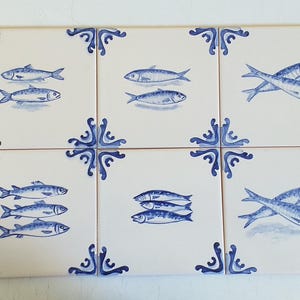Op de afbeelding: Een set van zes vierkante keramische tegels gerangschikt in een raster, elk met een witte achtergrond met blauwe decoratieve hoekversieringen en een centrale illustratie van vissen. De visontwerpen variëren per tegel, met enkele, gestapelde of gekruiste vissen.