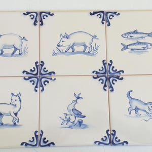 Op de afbeelding: Een set van zes vierkante keramische tegels gerangschikt in een raster, met witte achtergronden en handgeschilderde blauwe illustraties. Elke tegel toont een uniek diermotief, waaronder varkens, vissen, een vos, een vogel en een kat, omlijst door blauwe hoekaccenten.