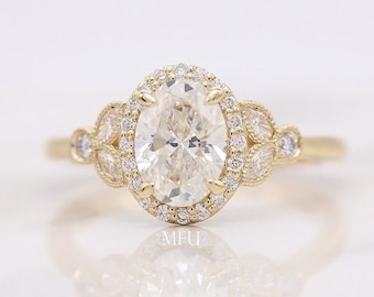 May include: A gold engagement ring with an oval-shaped center diamond surrounded by a halo of smaller diamonds. The band features a delicate design with smaller diamonds set in a leaf pattern.