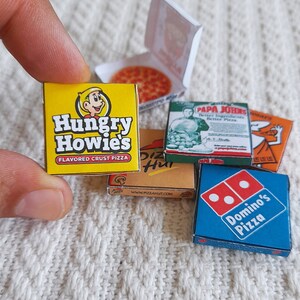 May include: A collection of miniature pizza boxes featuring popular pizza chains. The boxes are yellow, blue, green, and brown with the logos of Hungry Howie's, Pizza Hut, Papa John's, and Domino's Pizza.