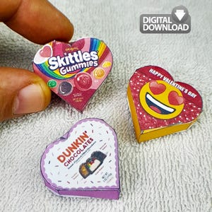 Może przedstawiać: Trzy pudełka w kształcie serca: jedno z opakowaniem Skittles Gummies, drugie z uśmiechniętą emotikoną i napisem "Happy Valentine's Day", a trzecie z opakowaniem Dunkin' Chocolates. Pudełka są kolorowe i wydają się być miniaturowymi wersjami.