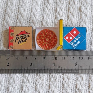 May include: Two miniature pizza boxes, one from Pizza Hut and one from Domino's Pizza, are placed side-by-side on a ruler. The Pizza Hut box is brown with a red and white logo. The Domino's Pizza box is blue with a red and white logo. The ruler is marked in inches.