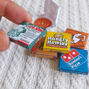 May include: A collection of miniature pizza boxes from different pizza chains. The boxes are arranged on a white knitted surface. The boxes are from Papa John's, Pizza Hut, Hungry Howie's, Little Caesars, and Domino's.