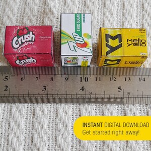May include: Three miniature cardboard boxes of popular soda brands. The boxes are red, green, and yellow. The red box says "Crush Strawberry" and has a strawberry on it. The green box says "7 Up Zero Sugar" and has a 7 Up can on it. The yellow box says " Mello Yello" and has a Mello Yello can on it. The boxes are lined up on a ruler with the numbers 5, 6, 7, 8, 9, 10, 11, 12, 13, and 14 visible.