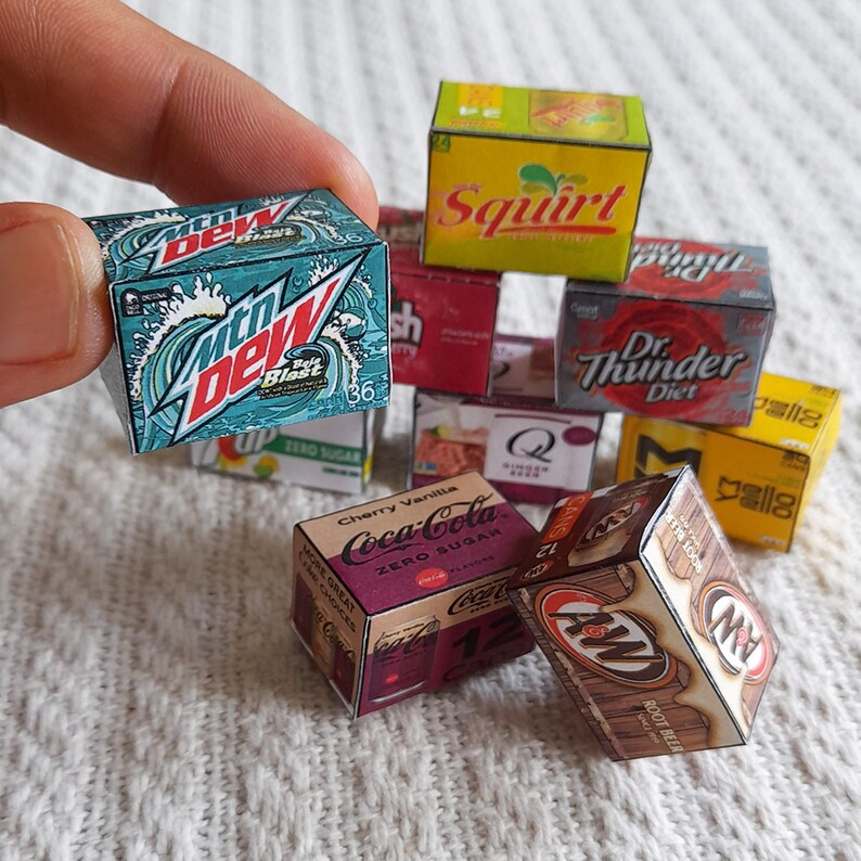 May include: A collection of miniature cardboard boxes featuring various soda brands, including Mountain Dew, Coca-Cola, Dr. Thunder, Squirt, and A&W Root Beer. The boxes are stacked and arranged on a white fluffy surface.