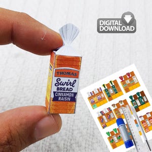 May include: A miniature paper model of a Thomas Swirl Bread Cinnamon Raisin loaf, held between fingers. The packaging is orange and yellow with the brand name and product details. A "DIGITAL DOWNLOAD" icon is in the background.