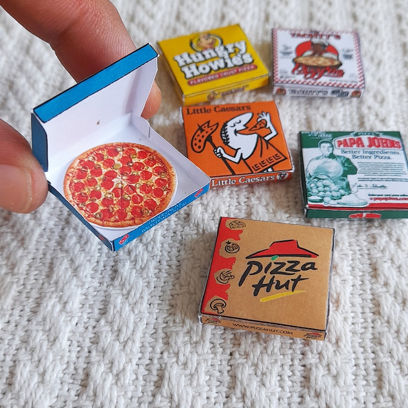 May include: A collection of miniature pizza boxes from different pizza chains. The boxes are arranged on a white fuzzy surface. The boxes are from Hungry Howies, Little Caesars, Papa Johns, and Pizza Hut.