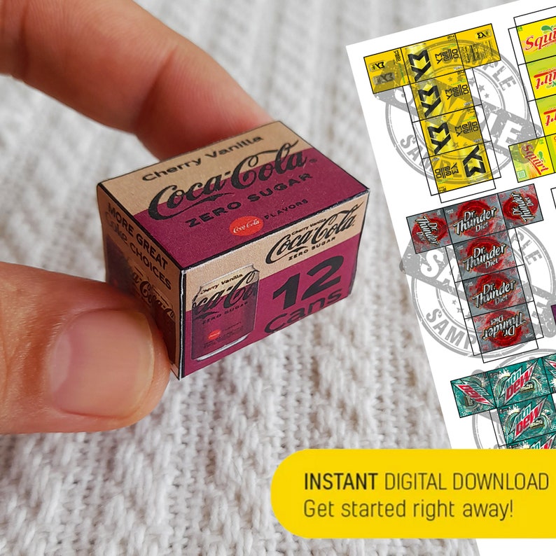 May include: A miniature cardboard box of Coca-Cola Cherry Vanilla Zero Sugar soda. The box is red and white with a picture of a can on the front. The box says "Cherry Vanilla" and "Zero Sugar" on the front. The box also says "12 Cans" on the front. The box is being held by a hand.