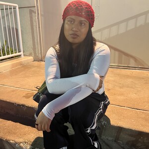 May include: A person wearing a red crochet hat, a white long-sleeved shirt, and black pants with white stripes. They are sitting on a set of concrete steps.