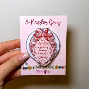Puede incluir: Un agarre circular para lector electrónico sobre una tarjeta de soporte rosa claro. El agarre presenta un marco festoneado rojo y blanco con un lazo rojo en la parte superior y el texto 'girls just wanna read books' en rojo. La tarjeta muestra 'E-Reader Grip' y 'Decifra'.