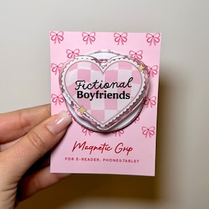May include: A heart-shaped magnetic grip for e-readers, phones, and tablets, featuring a pink and white checkered pattern with the text Fictional Boyfriends. The grip is mounted on a pink display card decorated with a repeating bow pattern.