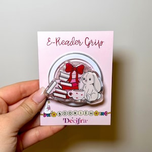 Può includere: Un'impugnatura rosa per e-reader con una base circolare trasparente. L'impugnatura presenta un'illustrazione a cartoni animati di una pila di libri rossi, un fiocco rosso, un coniglietto bianco e una tazza da tè. È visibile anche il testo "E-Reader Grip" e "Bookish Home".