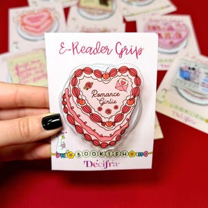 Puede incluir: Un agarre para lector electrónico con diseño de pastel en forma de corazón con glaseado rosa, cerezas rojas y rosas. El texto dice Romance Girlie. La tarjeta de respaldo muestra el texto E-Reader Grip, BOOKISH y Decifra, con una ilustración de un ganso.