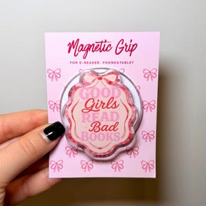 Könnte beinhalten: Ein pinkfarbener Magnetgriff für E-Reader, Telefone und Tablets. Der Griff zeigt den Text "GOOD Girls READ Bad BOOKS" in Pink und Rot. Die Verpackung ist rosa mit Schleifenmotiven.