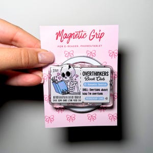 Puede incluir: Un accesorio rectangular Magnetic Grip para lectores electrónicos, teléfonos y tabletas con un gráfico de un esqueleto leyendo un libro. El diseño se asemeja a una tarjeta de membresía del Overthinkers Book Club con texto, código de barras y corazones rosas.