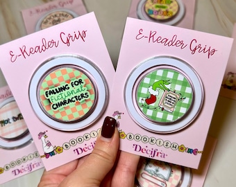 May include: Two circular e-reader grips on pink backing cards. The left grip has a green and orange checkerboard pattern with the text 'FALLING FOR FICTIONAL CHARACTERS'. The right grip features a green and white gingham pattern with a green hand.