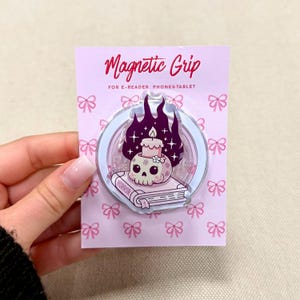 May include: A circular Magnetic Grip for e-readers, phones, and tablets on a light pink backing card with a bow pattern. The grip features a graphic of a light pink skull with a lit candle on top, resting on a book labeled HORROR with dark flames.