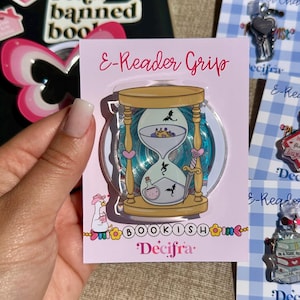 Può includere: Un accessorio E-Reader Grip su un cartoncino espositore rosa chiaro. L'impugnatura presenta l'illustrazione di una clessidra color sabbia contenente una corona, una pozione e piccole sagome di draghi neri. Una spada argentata è fissata sul lato.
