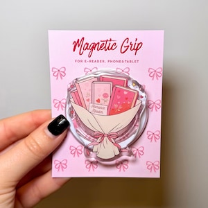May include: A circular magnetic grip for e-readers, phones, and tablets on a light pink backing card with a bow pattern. The grip features an illustration of a bouquet of pink books with the text Romance Reader, tied with a pink ribbon.