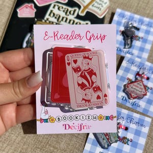 Peut inclure: Un support de liseuse rose avec un motif de carte à jouer représentant une reine tenant un livre, intitulé "Queen of Books". Le support est emballé avec d'autres breloques et accessoires, dont une clé et une breloque avec l'inscription "this holds my fictional boyfriend."
