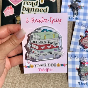 Può includere: Un'impugnatura per e-reader in acrilico con una pila di libri colorati e il testo 'IN A TOXIC RELATIONSHIP with my TBR' stampato sul davanti. Il design include copertine pastello, motivi a cuore e piccoli accenti a forma di stella.