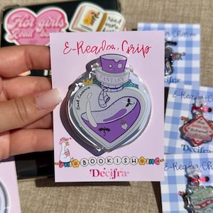 Può includere: Una spilla trasparente a forma di cuore con un design a pozione viola, con la parola "FANTASY" e il testo "Book Essence". La spilla include un fantasma, draghi e il testo "E-Reader Grip" e "BOOKISHOME Decifra."
